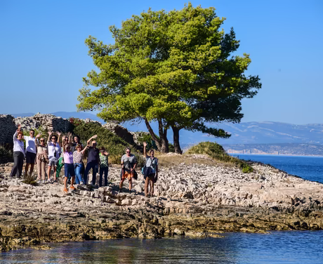 Eine Gruppe voller Menschen stehen am Steinigen Ufer und winken in die Kamera